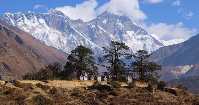 Aerial shot of white chortens with colorful prayer flags set in green forest with Ama Dablam and surrounding Everest Himalayan peaks rising beneath a cloudy blue sky in Nepal.