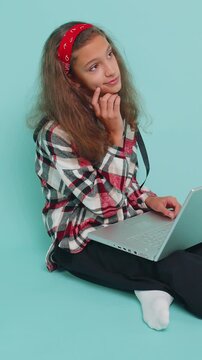 Happy young teenager child girl kid 12 years old works on notebook laptop pc, sends messages, makes online purchases, watching movies. Preteen children isolated sitting on studio blue background