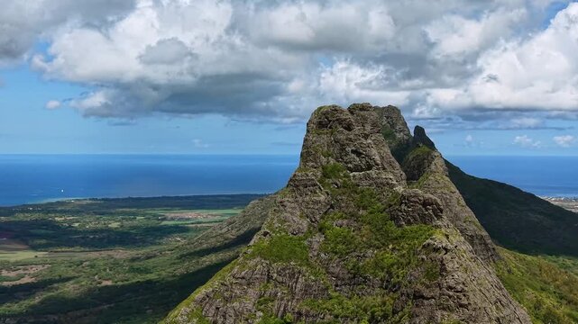 First keyframe: Mountain peaks frame a vast ocean view. Partly cloudy sky looms overhead. Lush green slopes extend toward the sea. Second keyframe: Close-up shows rugged rock textures