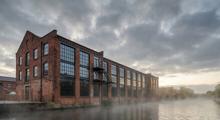 Fototapeta premium A brick industrial building with large windows stands beside a misty canal under a cloudy sky.