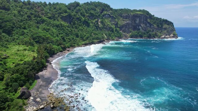 Aerial drone footage of Pangasan beach from the eastern rice field point of view, with black sand beach, cliffs, blue sea, waves, in a sunny weather, in Pacitan regency, Java island, Indonesia