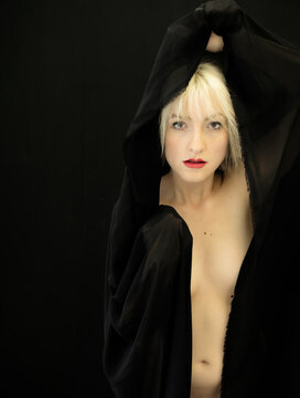 Blonde woman framed by flowing black fabric raises her arms above head revealing face and red lips with direct gaze against deep dark background creating tension and intrigue