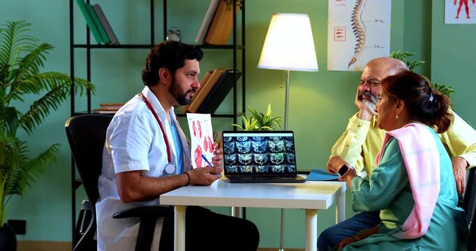 Professional Indian young male doctor explaining organ functions using a graphic figure to Asian senior retired couple patients across a table in a modern clinic; healthcare education concept.