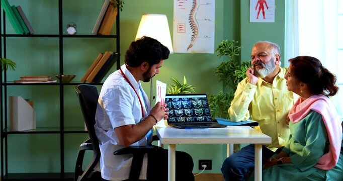 Professional Indian young male doctor explaining organ functions using a graphic figure to Asian senior retired couple patients across a table in a modern clinic; healthcare education concept.