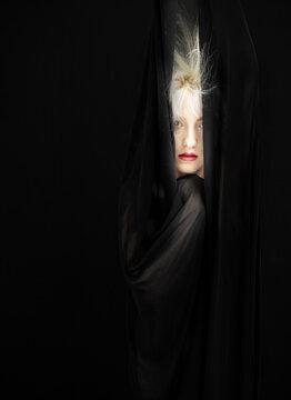 Blonde woman peers through dark flowing fabric revealing only part of her face with striking red lips dramatic contrast and mystery against deep black background