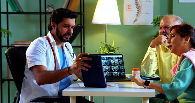 Professional Indian young male doctor using touchscreen tablet computer with Asian senior retired couple patients across a table in a modern clinic; healthcare, technology, and old age concept.