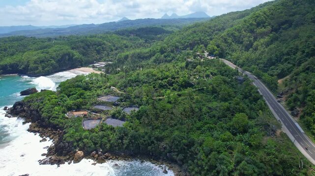 Aerial drone footage of Watu Papak beach bay, in Ngadirojo area, Pacitan regency , Java island, Indonesia, the main road passing by thru green mountains, some food stalls, and fish farm ponds aside