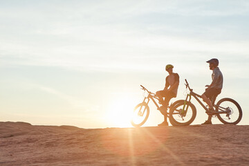 Fototapeta premium Men, mountain bike or rest with sunset in desert for extreme sport or fitness together in nature. Flare, male people or cyclist with friend or bicycle for cycling break or outdoor challenge on space