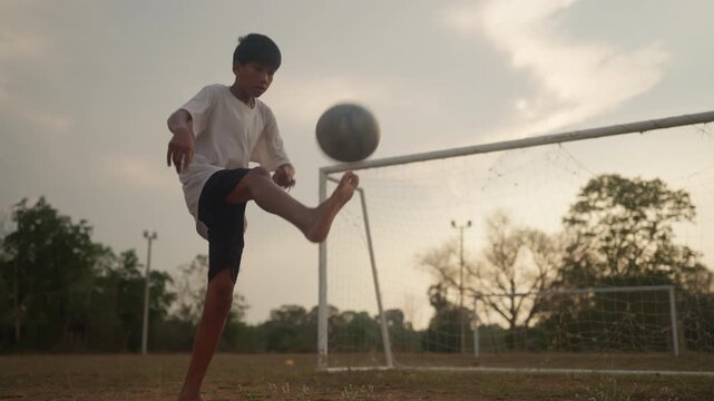 Young boy kicks soccer ball toward goal. Sunset sky casts warm glow over field. Green grass, trees, and goalpost visible. Child wears white shirt and dark shorts. Ball arcs toward net in open space
