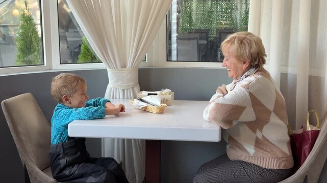 An elderly woman and a young boy sit at a cafe table, smiling warmly at each other. They share a happy, connected moment in a bright, cozy room. Their joy makes the atmosphere feel very sweet.