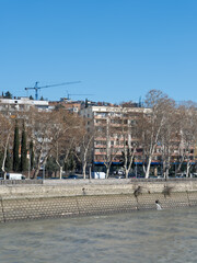 View of the stone embankment along the Mtkvari River in Tbilisi, Georgia, featuring classic residential apartments and leafless trees under a clear blue sky during the winter season. © Microscope