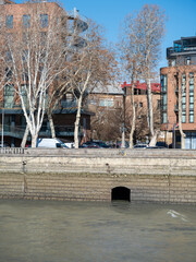 View of the stone embankment along the Mtkvari River in Tbilisi, Georgia, featuring classic residential apartments and leafless trees under a clear blue sky during the winter season. © Microscope
