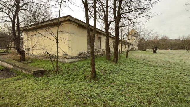 An old, long building rests behind a row of winter trees. The frost-covered grass and grey sky create a lonely, quiet mood. This peaceful scene feels like a cold morning frozen in time.