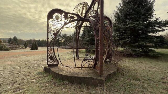 A decorative metal pavilion stands on a stone base facing a wide park path. The frosted grass and soft morning light create a serene and magical feeling in the quiet garden.