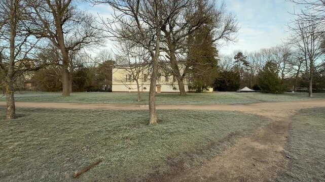 The historic yellow mansion stands quietly behind a field of silver frost. Bare winter trees and a winding dirt path create a nostalgic, peaceful feeling in the cold morning air.