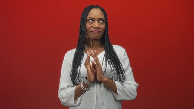 Woman with hands clasped in prayer, white blouse, bracelets and smartwatch in studio with red backdrop; serenity reflection.
