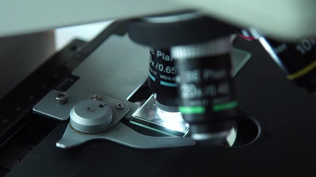 Close-up of a microscope examining a glass slide with a gloved hand in a laboratory, illustrating scientific research and microscopic analysis.