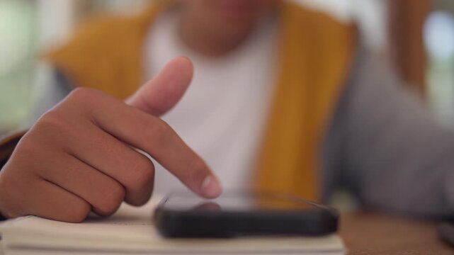 Close-up hand taps smartphone screen. Finger moves slowly over phone surface. Person wears yellow vest, white shirt. Background softly blurred, out of focus. Hand rests near stack of white books