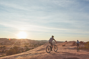 Fototapeta premium Men, mountain bike or cycling with sunset in desert or dunes for extreme challenge or sport. Active, male people or cyclist with bicycle for outdoor trip or travel on dirt track together in nature