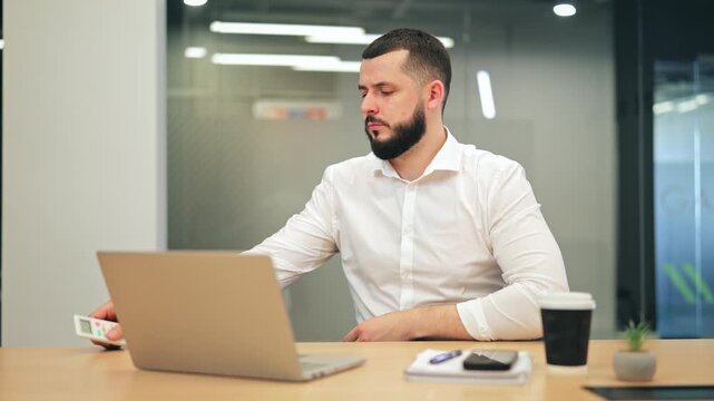 Feeling cold in office, professional adjusts air conditioning with remote control, then resumes work on laptop. Focused, determined to achieve comfort, productivity in modern workspace.