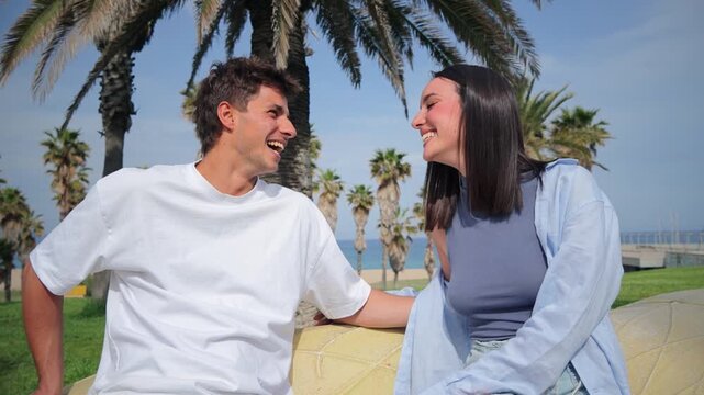 Delighted young couple enjoying playful banter and genuine smiles while seated comfortably outdoors near palm trees.