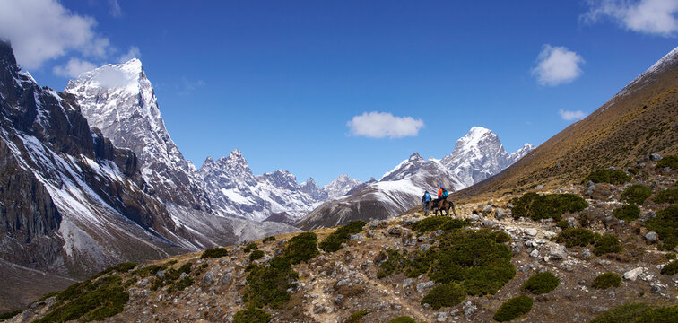 Breathtaking wide panoramic view of snow-capped Himalayan range under bright sunny sky, with two trekkers on Everest Base Camp trail - one riding horse with backpack high mountain path 