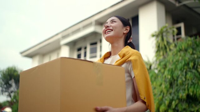 Happy young woman holding moving box and house keys in front of new home. Moving day, real estate purchase, mortgage loan, home ownership and property investment concept.
