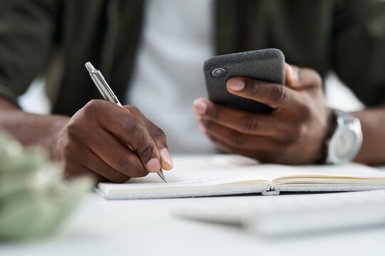 Phone, writing and hands of businessman with notebook in office for agenda, planning or contact. Technology, journal and male creative editor with diary and cellphone for feedback on publishing.