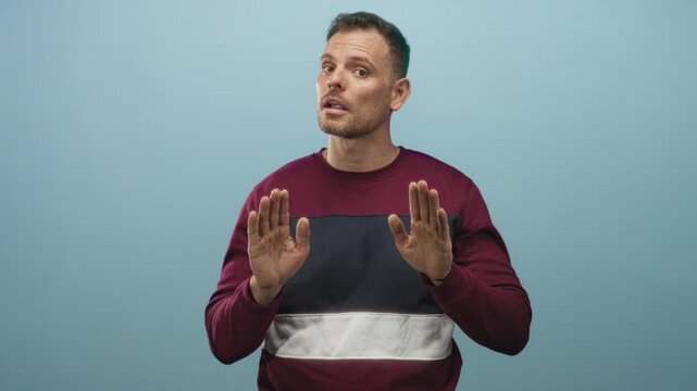 Man young with hands forward palms out stopping gesture, wearing maroon and navy sweatshirt, looking wary in a studio; hesitation.
