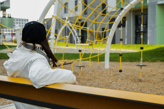 Young woman in a white jacket sits quietly on a bench, looking toward a modern playground with bright climbing equipment.