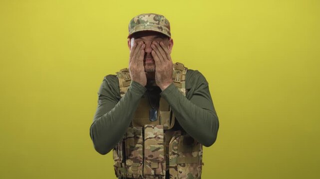 Man soldier in camouflage vest and cap with dog tags clenches fists, squints and rubs his eyes in studio; fatigue.
