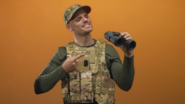 Man soldier in camouflage uniform points finger to binoculars in studio wearing tactical vest and dogtags; confidence duty.