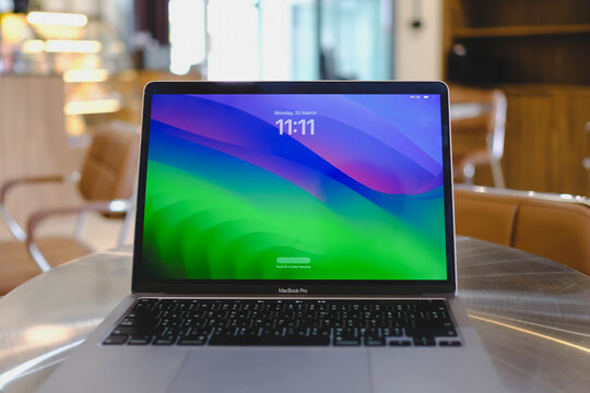 Bangkok, Thailand. March 30, 2026; A MacBook Pro laptop displaying a colorful lock screen with the date and time is placed on a round metallic table in a modern cafe.