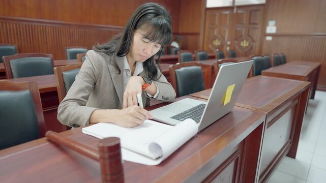 Woman studies law in formal courtroom setting. She writes notes, uses laptop beside her. Wooden benches, polished tables, official ambiance. Focused, serious expression, professional demeanor