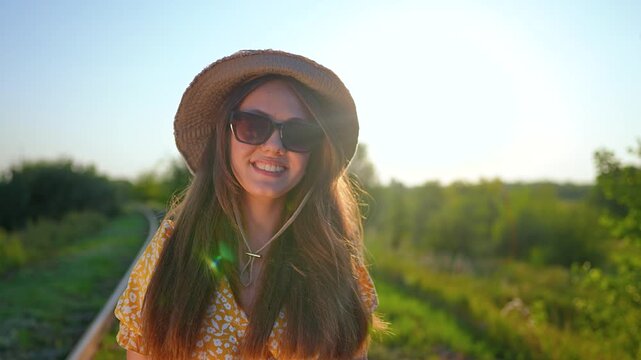 Smiling woman in hat and sunglasses near railway track at sunset. Happy female with long hair by railroad in countryside. Woman smiling in dress near train rail at golden hour outdoors.