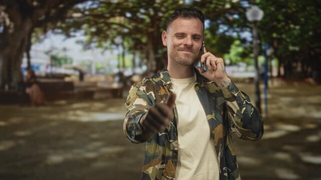 Man in camouflage jacket on smartphone giving thumbs up and pointing gesture while smiling in a sunlit park with trees, benches and lampposts; confidence.
