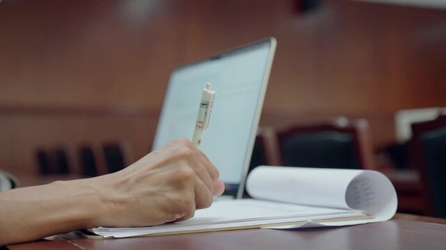 Hand writes notes beside open laptop in courtroom. Rolling document rests beside laptop on wooden table. Background shows courtroom seating and polished wood