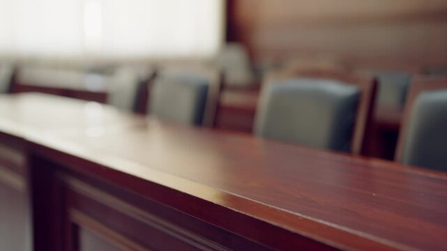 Empty courtroom with polished wood tables and dark chairs. Rows of chairs face long, gleaming tables. Soft focus highlights the quiet, formal atmosphere. Wooden surfaces reflect subdued