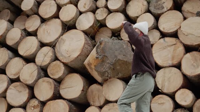 A woman walks along a path next to a large stack of logs in the forest. The natural setting features many trees and a wooden wall of circular cuts.