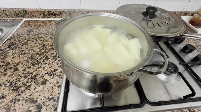 Cooking peeled cassava root in boiling water on a gas stove in a stainless steel pot