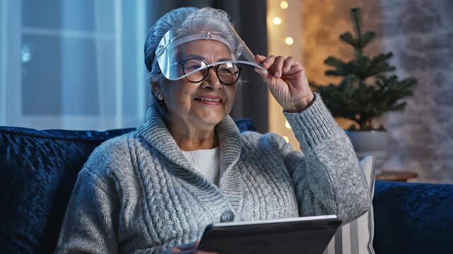 Senior woman wears shield while using tablet on sofa. Elderly woman with visor reads tablet at home. Senior protection with shield and tablet. Woman uses digital device wearing protective visor.