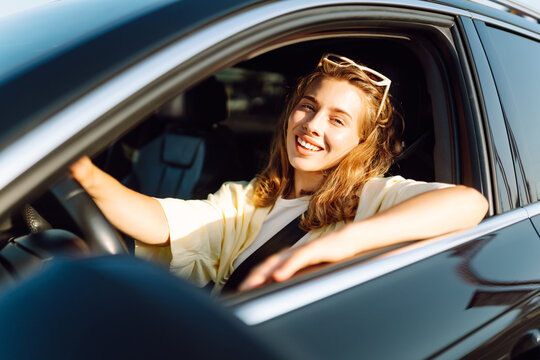 A young woman sits behind the wheel of a car and enjoys the sunny day. A smiling female driver looks out the window at sunset. Concept of transportation, city, and leisure.