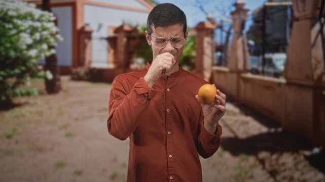 Man holds orange, touches throat and winces outside a building on a sunlit walkway; discomfort allergy reaction.