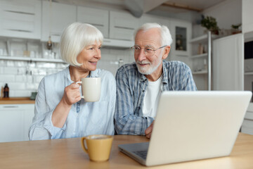 Fototapeta premium Senior couple drinking coffee at kitchen table planning travel on a laptop, medium close-up, bright scandinavian interior, morning natural light, candid interaction