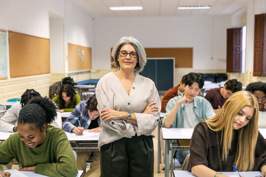 Senior teacher smiling, standing with arms crossed, observing multicultural high school or college students writing an exam in a bright classroom