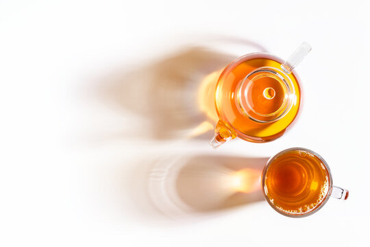 Top view of glass cup of tea and teapot with long shadows on white background.