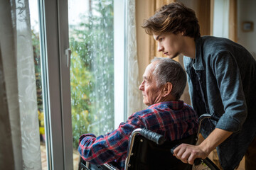 Obraz premium Adult son assisting elderly father in wheelchair near window, medium close-up, bright home interior, emotional family support moment simple design