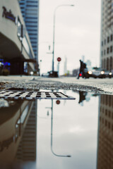 Obraz premium City street with puddle reflecting buildings and a person walking on a cloudy day in an urban setting