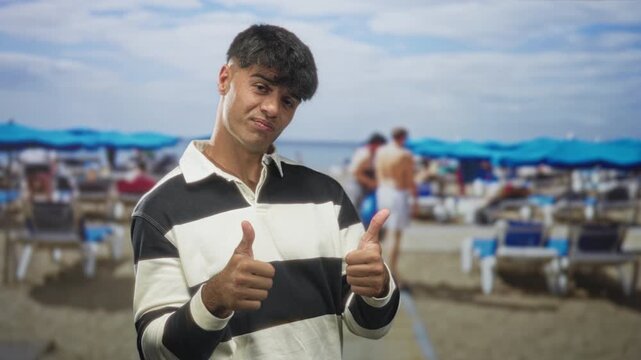 Young hispanic man in striped rugby shirt giving double thumbs up and pointing at camera on crowded beach with umbrellas and lounge chairs; playful confidence.