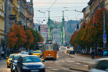 Obraz premium Yellow retro tram riding through the autumn street of Budapest toward Liberty Bridge with red trees.